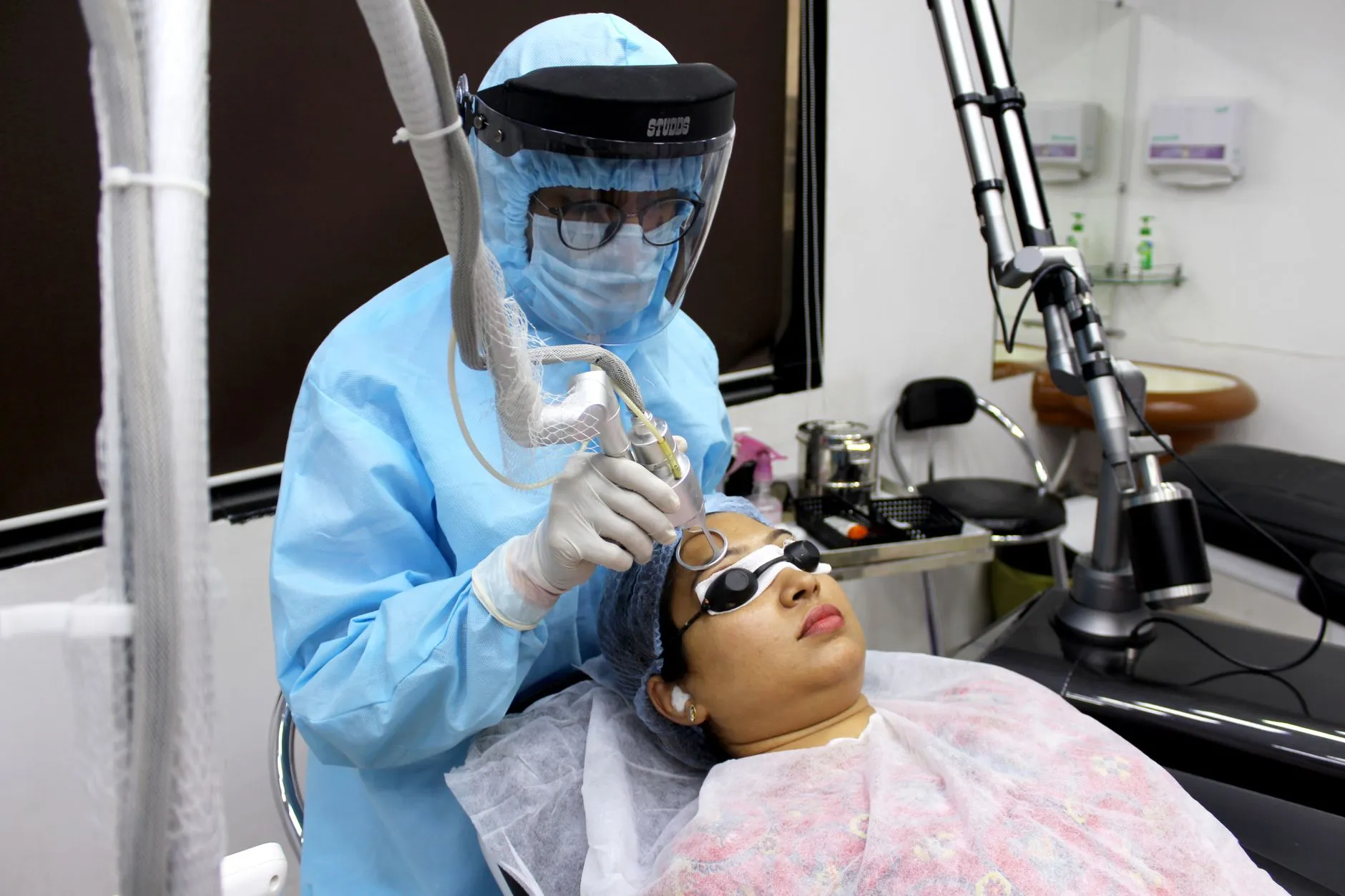 Nhân viên y tế mặc đồ bảo hộ thực hiện buổi chiếu laser trên vùng da mặt, người bệnh đeo kính che mắt.