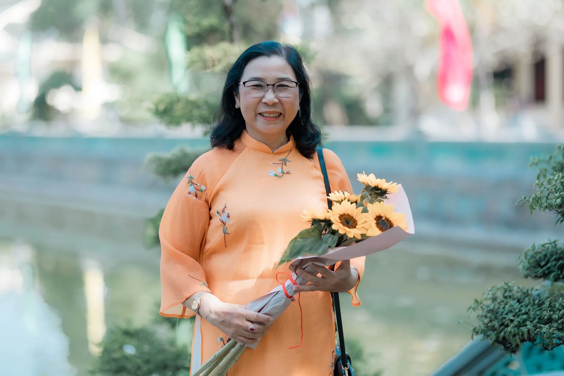 Phụ nữ trung niên mặc áo dài cầm bó hoa hướng dương, chụp ngoài trời.