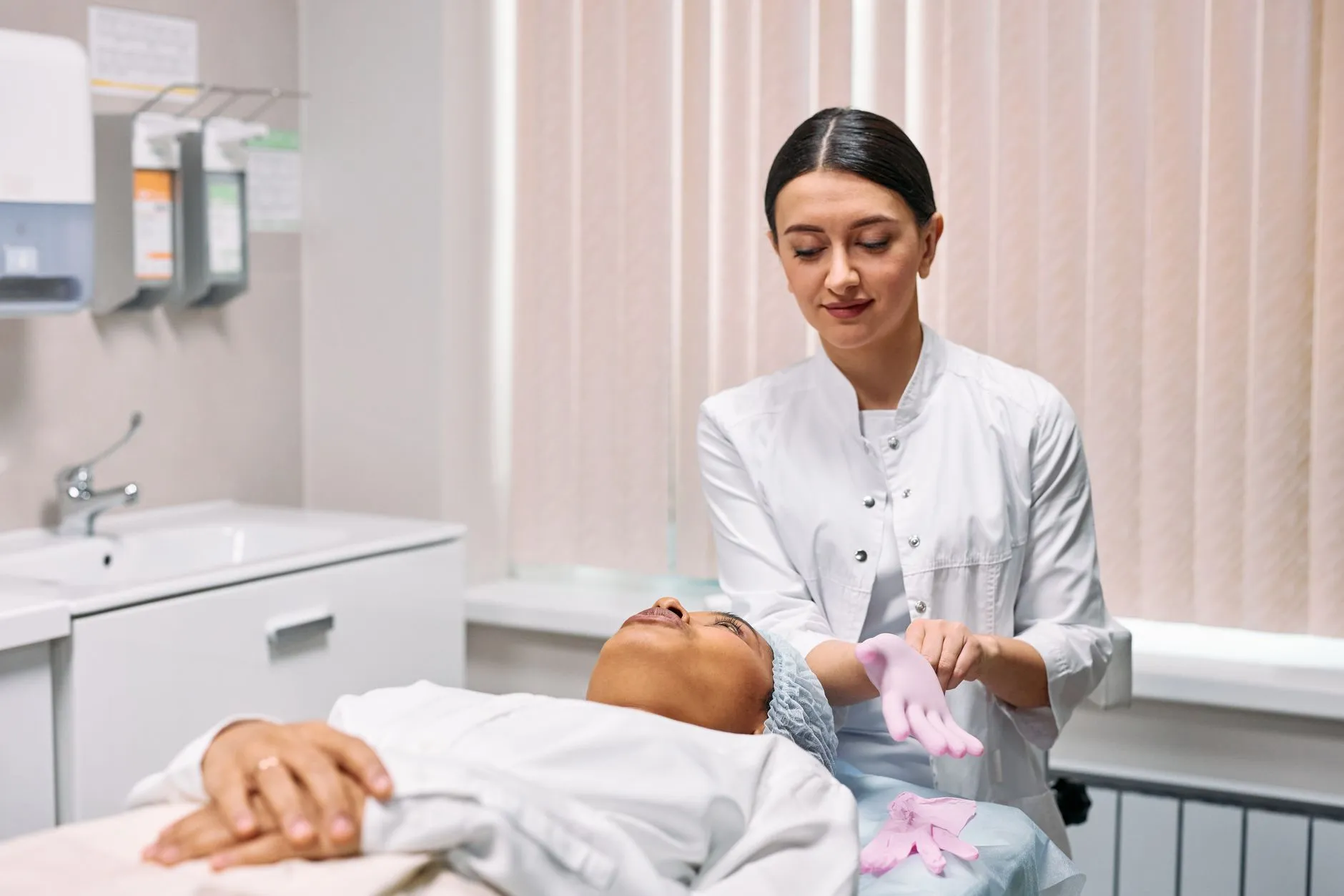Nhân viên y tế đeo găng tay bên giường thủ thuật, người bệnh nằm đội mũ phẫu thuật.