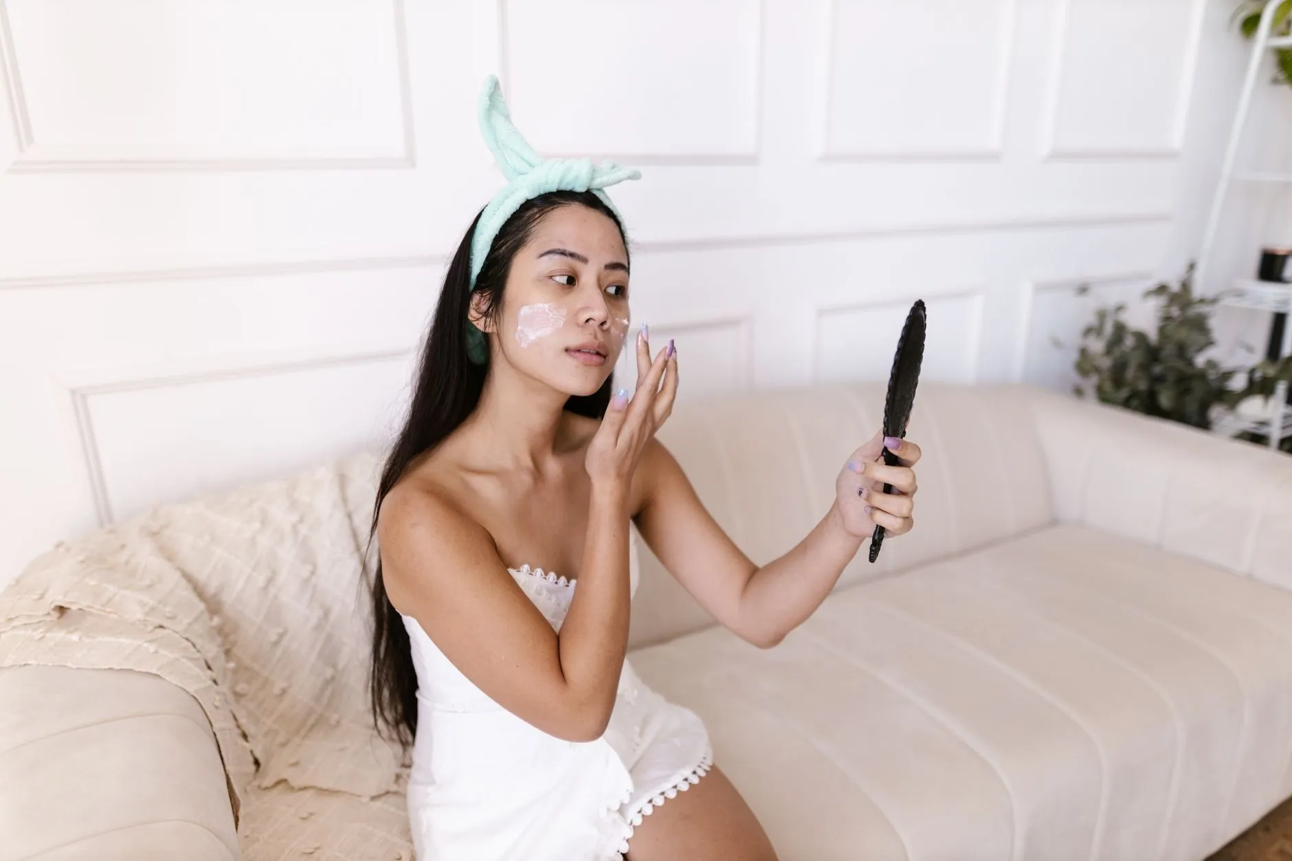Người phụ nữ cầm gương soi và thoa kem dưỡng lên vùng má.